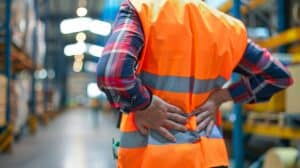 A warehouse worker in a safety vest holds his back in discomfort, highlighting the importance of workplace ergonomics. This image is ideal for industrial and safety themes.